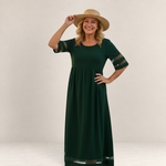 Valvet Oak House Penelope maxi dress in dark green with sheer sleeve details, worn by a smiling woman in a straw hat.