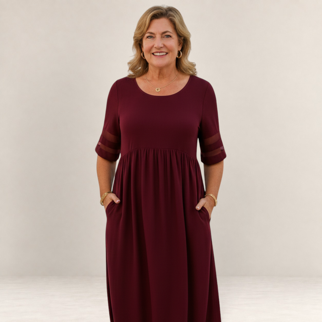 Valvet Oak House Penelope burgundy maxi dress with sheer sleeve details worn by a smiling woman in a coastal setting