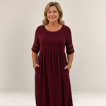 Valvet Oak House Penelope burgundy maxi dress with sheer sleeve details worn by a smiling woman in a coastal setting
