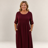 Valvet Oak House Penelope burgundy maxi dress with sheer sleeve details worn by a smiling woman in a coastal setting