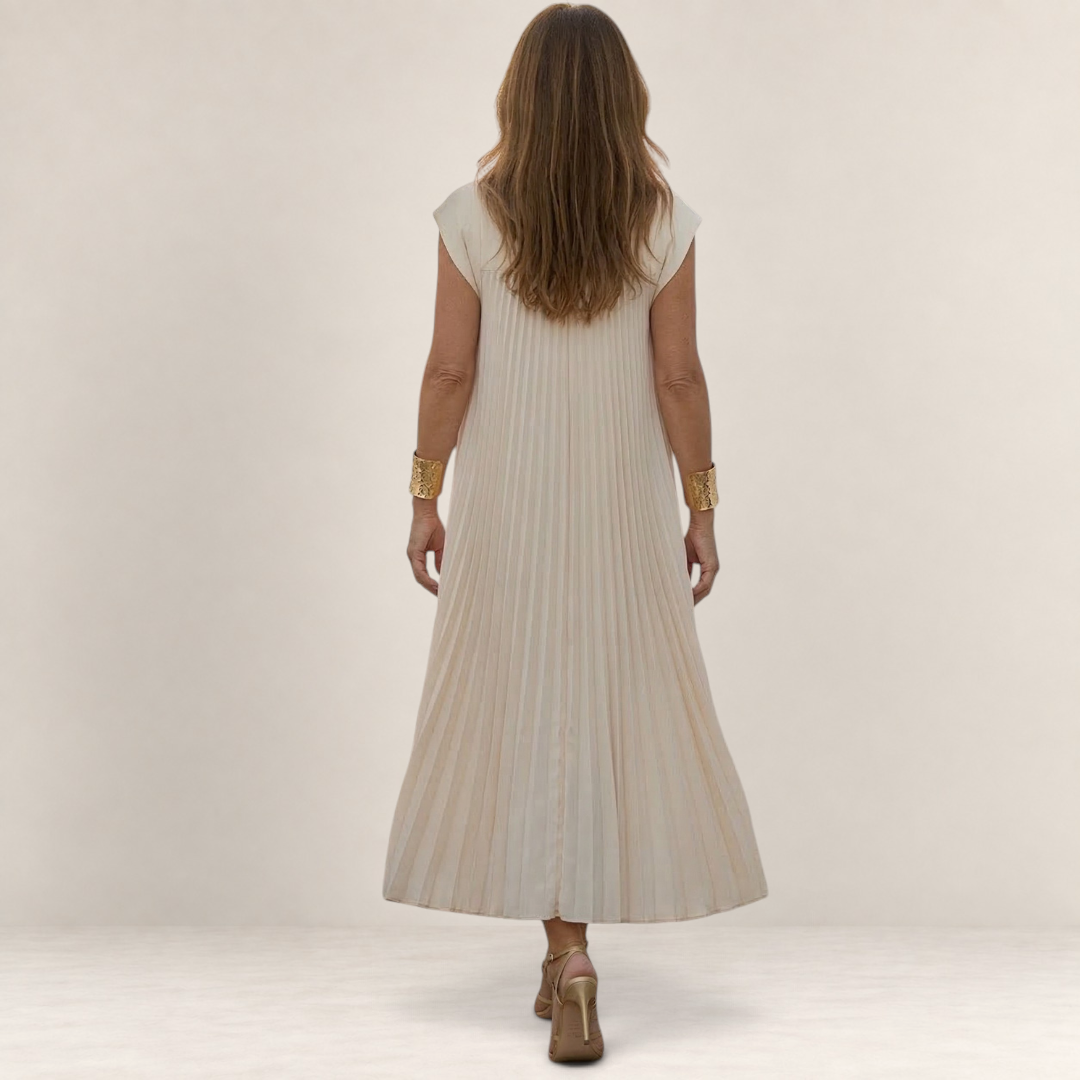 Valvet Oak House Chrysanthe cream pleated midi dress worn by a woman walking on a cobblestone street at sunset.