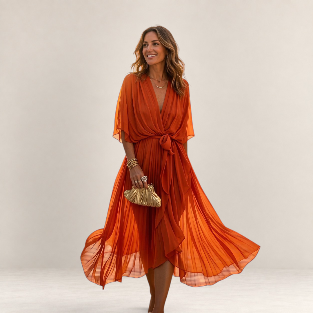 Valvet Oak House Helena orange pleated V-neck dress with a waist tie, worn by a woman in a Mediterranean village.