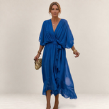 Valvet Oak House Helena blue flowy midi dress with V-neck and waist tie worn by a woman on a cobblestone street.