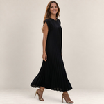 Valvet Oak House Chrysanthe black pleated midi dress worn by a woman on a cobblestone street at sunset