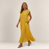 Valvet Oak House Chrysanthe yellow pleated maxi dress with flutter sleeves worn by a smiling woman in gold heels.