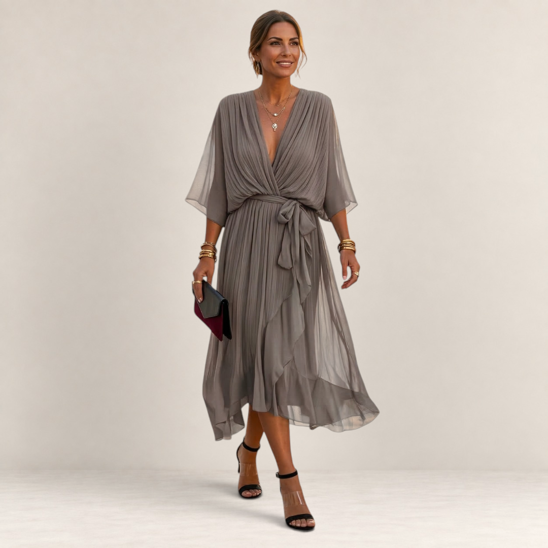 Valvet Oak House Helena grey pleated midi dress with V-neck and waist tie worn by a woman on a cobblestone street