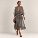 Valvet Oak House Helena grey pleated midi dress with V-neck and waist tie worn by a woman on a cobblestone street