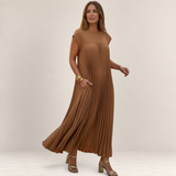Valvet Oak House Chrysanthe pleated maxi dress in tan worn by a woman on a cobblestone street at sunset.