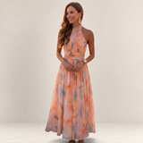 Valvet Oak House silk maxi dress with halter neckline in a peach floral print, worn by a woman at sunset in Greece.