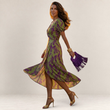 Valvet Oak House Lara floral wrap dress in green and purple, worn by a woman with a purple fringed handbag.