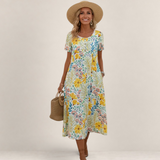 Valvet Oak House Margaret floral midi dress with short sleeves, worn by a smiling woman with a straw hat and bag.