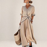 Valvet Oak House Mila beige midi dress with V-neck, contrast piping, and waist detail, worn by a woman by the sea.